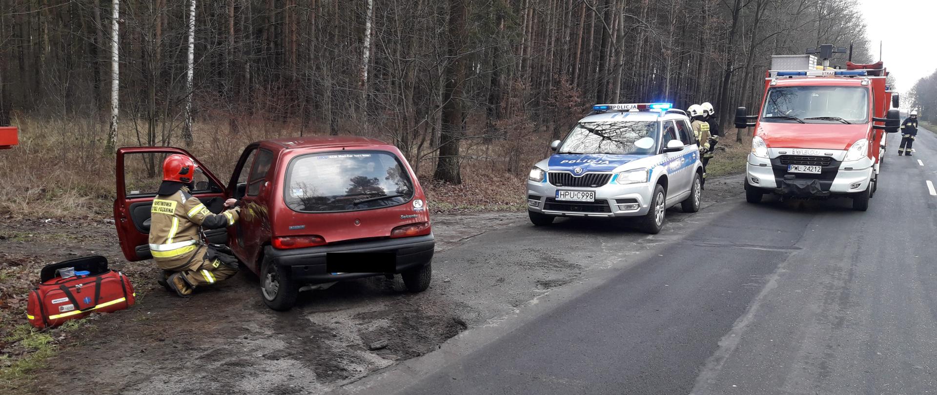 Rozbity samochód po prawej stronie. Strażak udziela pomocy. Z tyłu radiowóz policyjny oraz wóz strażacki.