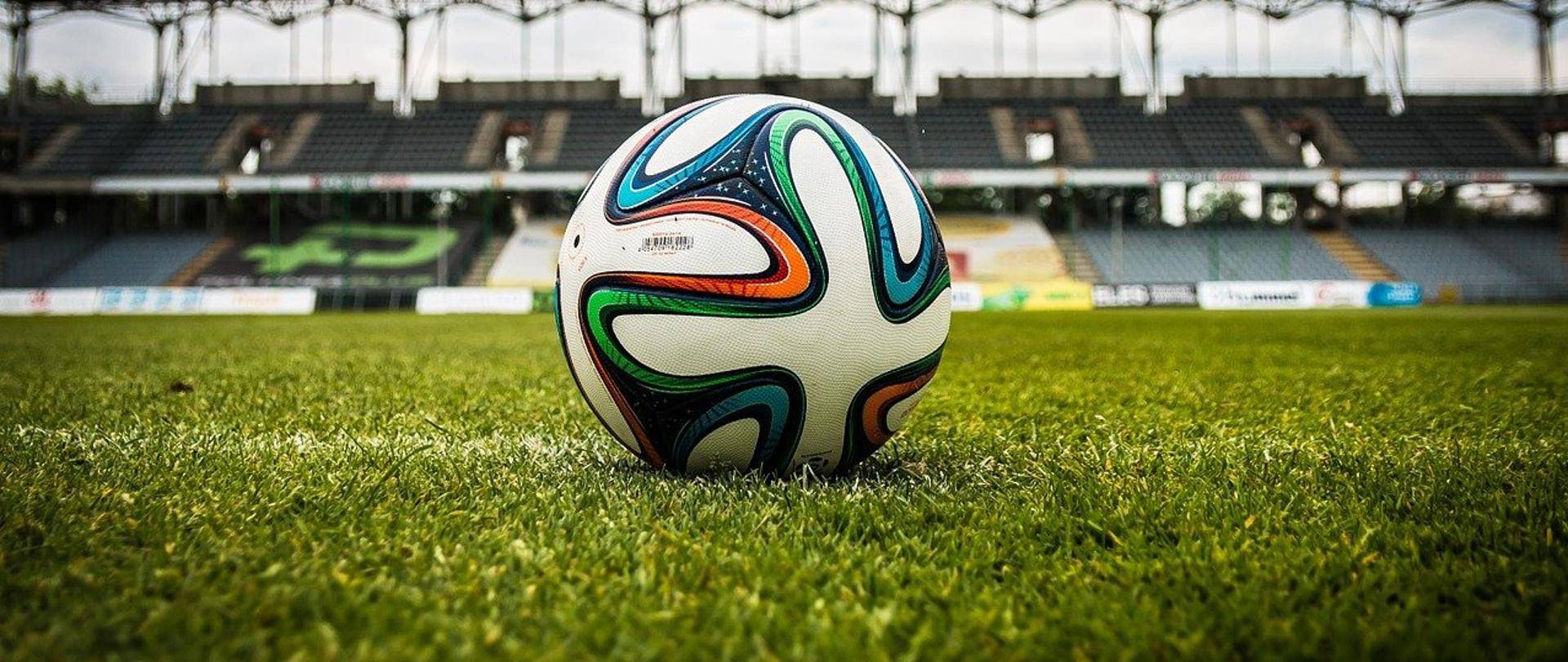 Zdjęcie przykładowe piłki nożnej - piłka na trawie. W tle trybuny stadionu. 