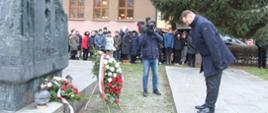 Minister Czarnek kłania się przed czarnym pomnikiem pod którym leżą wieńce biało-czerwonych kwiatów, w tle duża grupa ludzi.