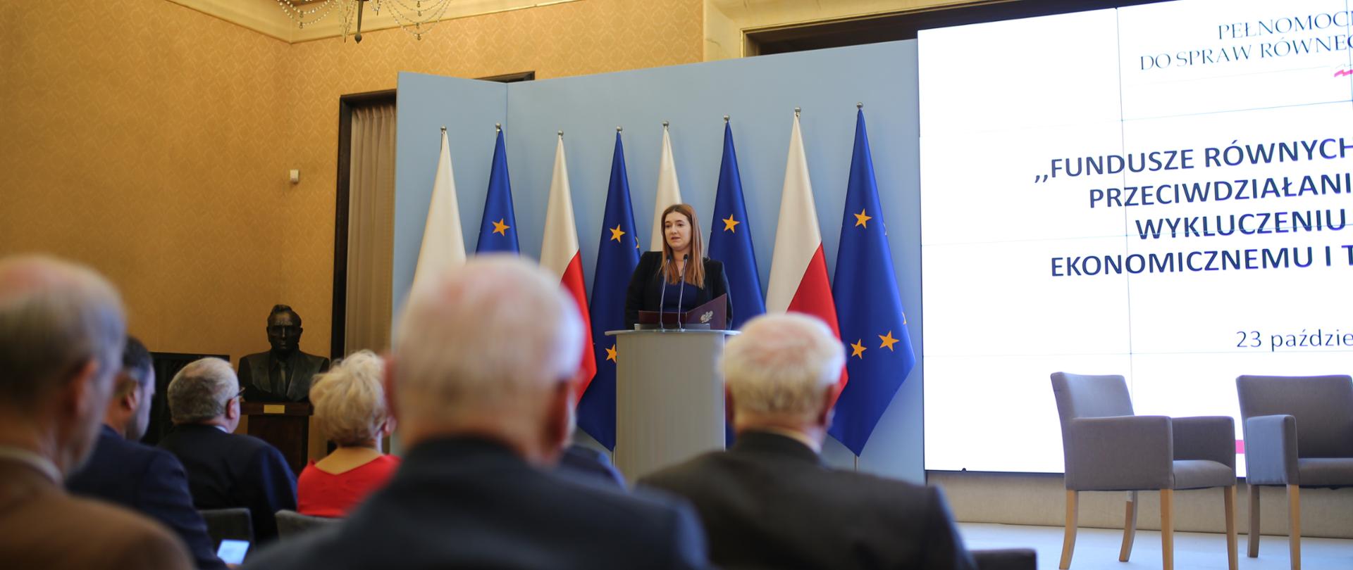 Na scenie w mównicy stoi wiceminister Anna Gembicka. Za nią flagi polskie i UE. Obok na ekranie wyświetla się napis: ""Fundusze równych szans sposobem na przeciwdziałanie dyskryminacji i wykluczeniu społecznemu, ekonomicznemu i technologicznemu". Przodem do sceny w rzędach siedzą na krzesłach uczestnicy konferencji.