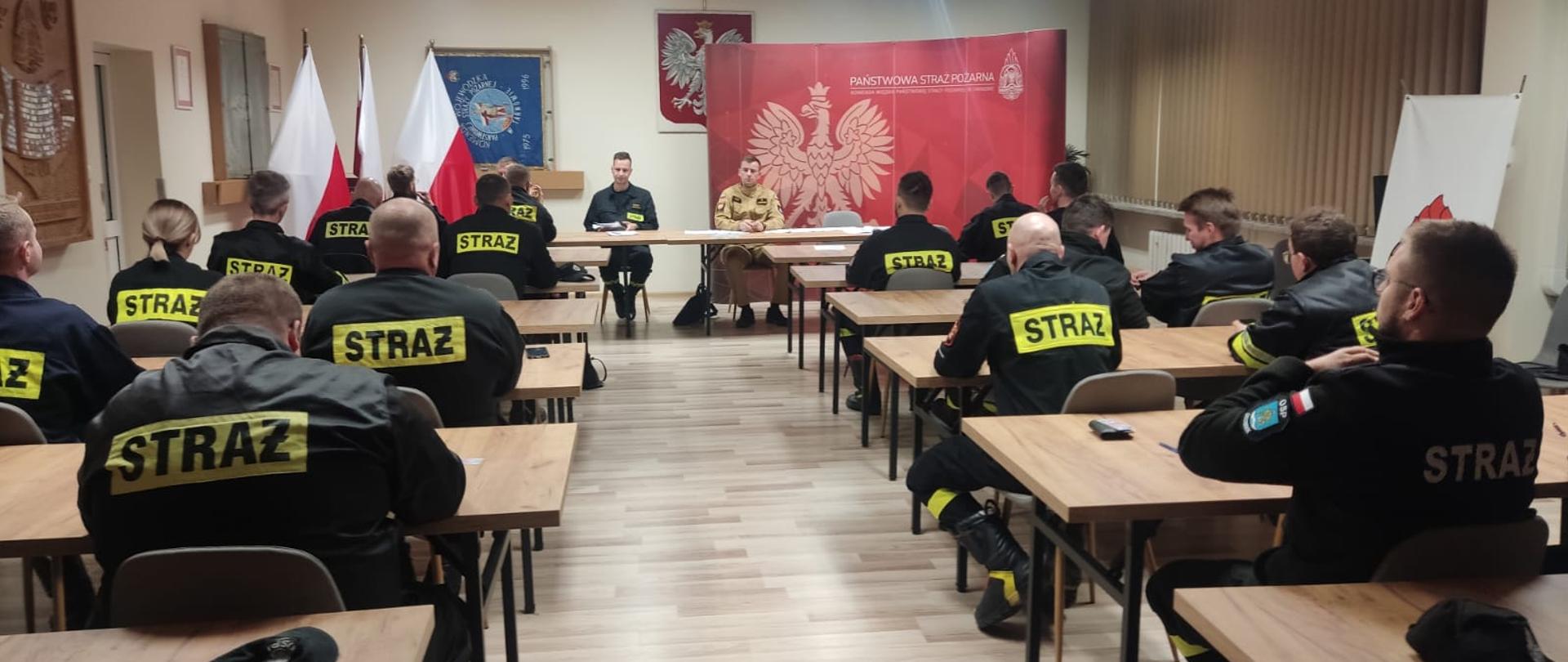 Strażacy ubrani w czarne ubrania koszarowe siedzą w ławkach przodem skierowani do stołu prezydialnego. Przy stole prezydialnym siedzą osoby prowadzące szkolenie, z tyłu godło Polski.