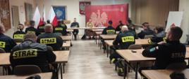 Strażacy ubrani w czarne ubrania koszarowe siedzą w ławkach przodem skierowani do stołu prezydialnego. Przy stole prezydialnym siedzą osoby prowadzące szkolenie, z tyłu godło Polski.