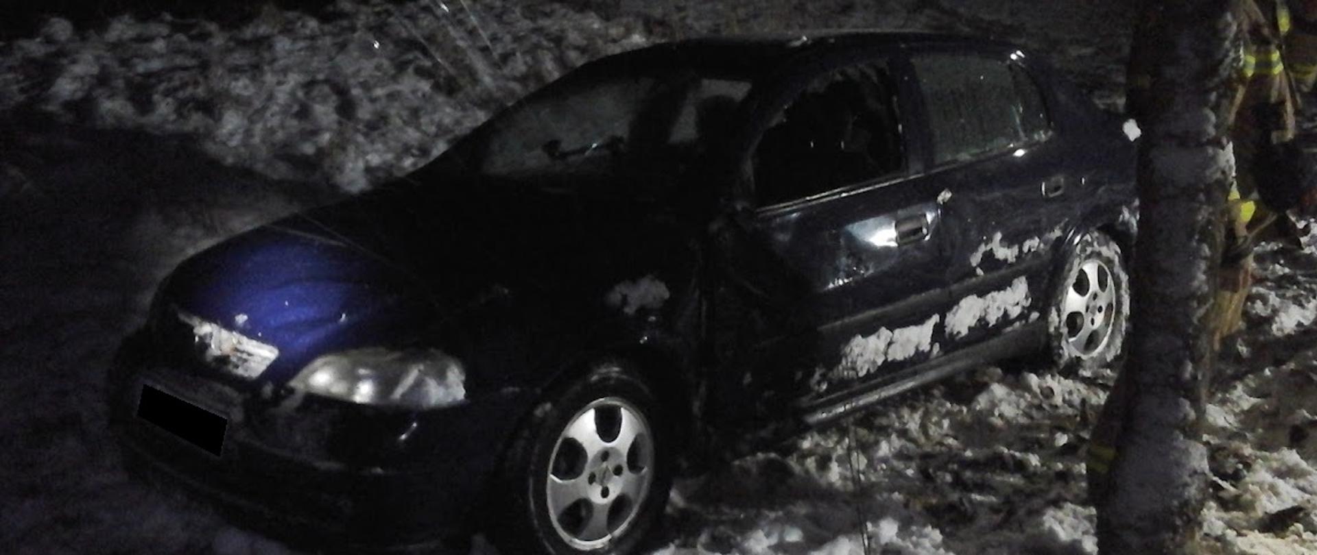 Samochód osobowy stoi na kołach na poboczu drogi. Wokół leży śnieg. Wybita szyba w drzwiach przednich od strony kierowcy. 