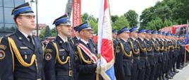 Rząd funkcjonariuszy PSP w umundurowaniu galowym z elementami złotego sznura i nakryciami głowy. Jeden ze strażaków trzyma sztandar z widocznym napisem „Służbie”. W tle sprzęt pożarniczy i baner KSRG.