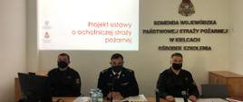 Przy stole prezydialnym siedzi trzech strażaków w mundurach i maseczkach ochronnych, w tle ekran z prezentacja dot. konsultacji ustawy o OSP 