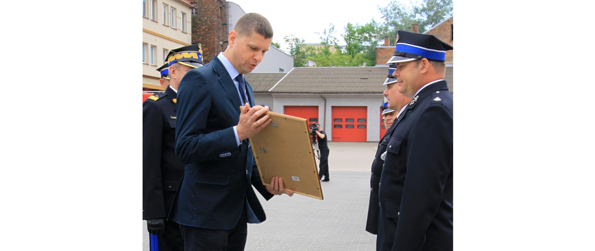 Na tle budynku garażowego z czerwonymi bramami wręczany jest akt strażakowi w granatowym mundurze. Wręczenia dokonuje Dariusz Piontkowski Wiceminister Edukacji i Nauki ubrany w granatowy garnitur.