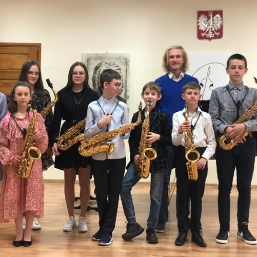 Siedmioro odświętnie ubranych dzieci, trzy dziewczynki i czterech chłopców stoi na scenie i trzyma złote saksofony. Obok nich stoi ich nauczyciel ubrany na niebiesko. W tle wisi godło. 