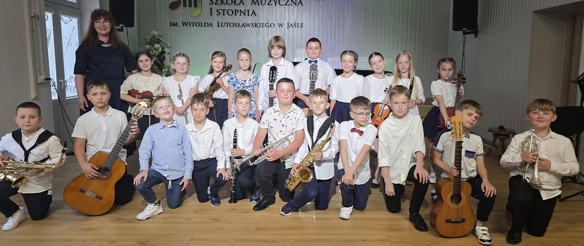 Zdjęcie przedstawiające dużą grupę uczniów oraz Panią Dyrektor PSM pozujących do zdjęcia na scenie auli PSM w Jaśle. Wszyscy ubrani w stroje galowe, trzymają swoje instrumenty i uśmiechają się. W tle logo PSM oraz godło, po bokach głośniki.