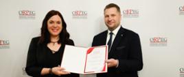 Minister przekazuje dyplom elegancko ubranej kobiecie, za nimi ściana z napisami ORPEG.