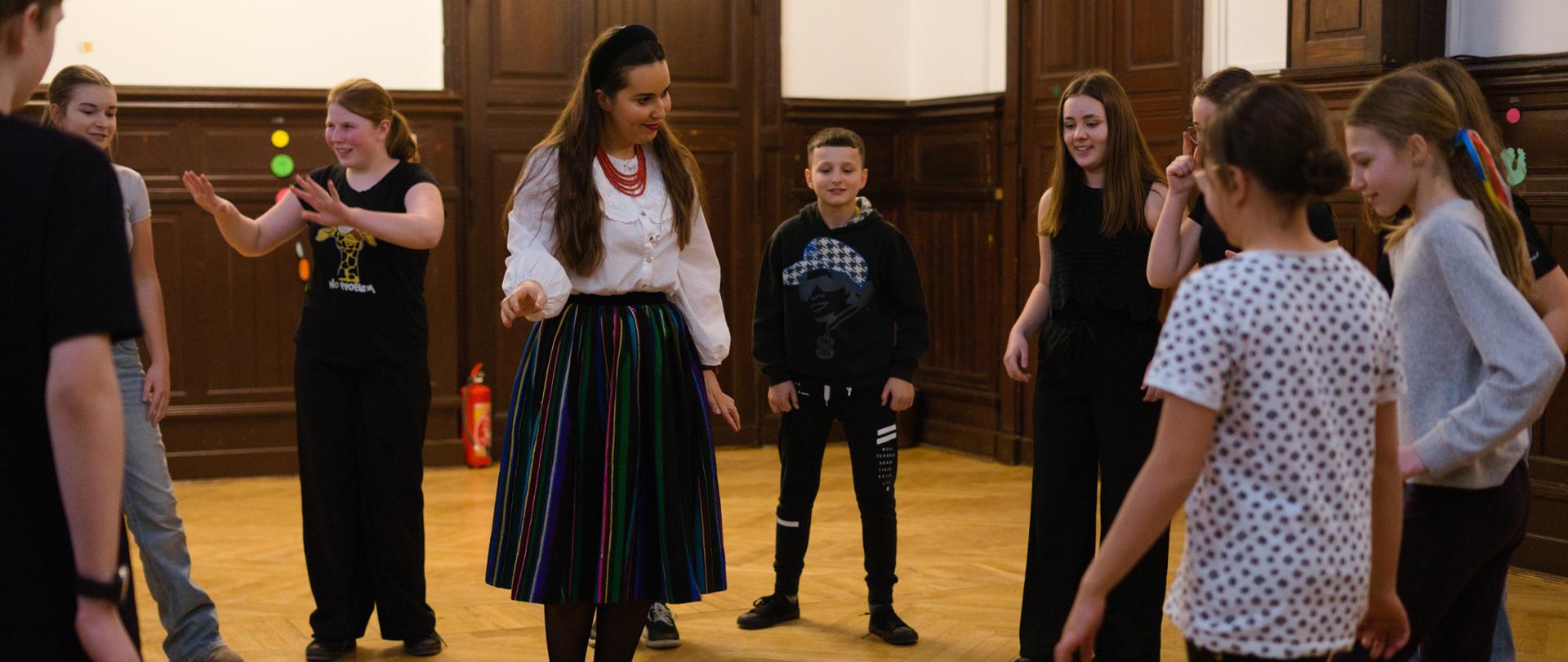 Grupa dzieci i młodzieży stoi w kręgu w przestronnej sali z drewnianą podłogą uczestnicząc w zajęciach ruchowych prowadzonych przez instruktorkę stojącą pośrodku. Na podłodze w centrum leży miotła upleciona z gałązek. 