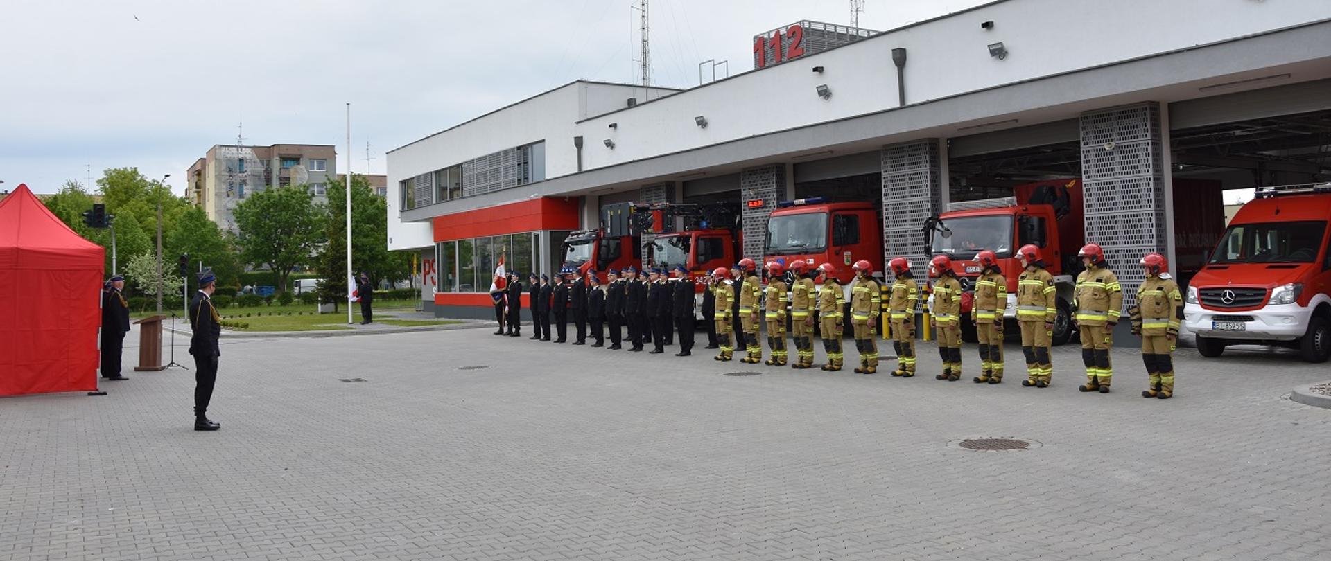 Na pierwszym planie ustawieni funkcjonariusze w rzędzie a za nimi wozy strażackie i budynek komendy Miejskiej PSP.