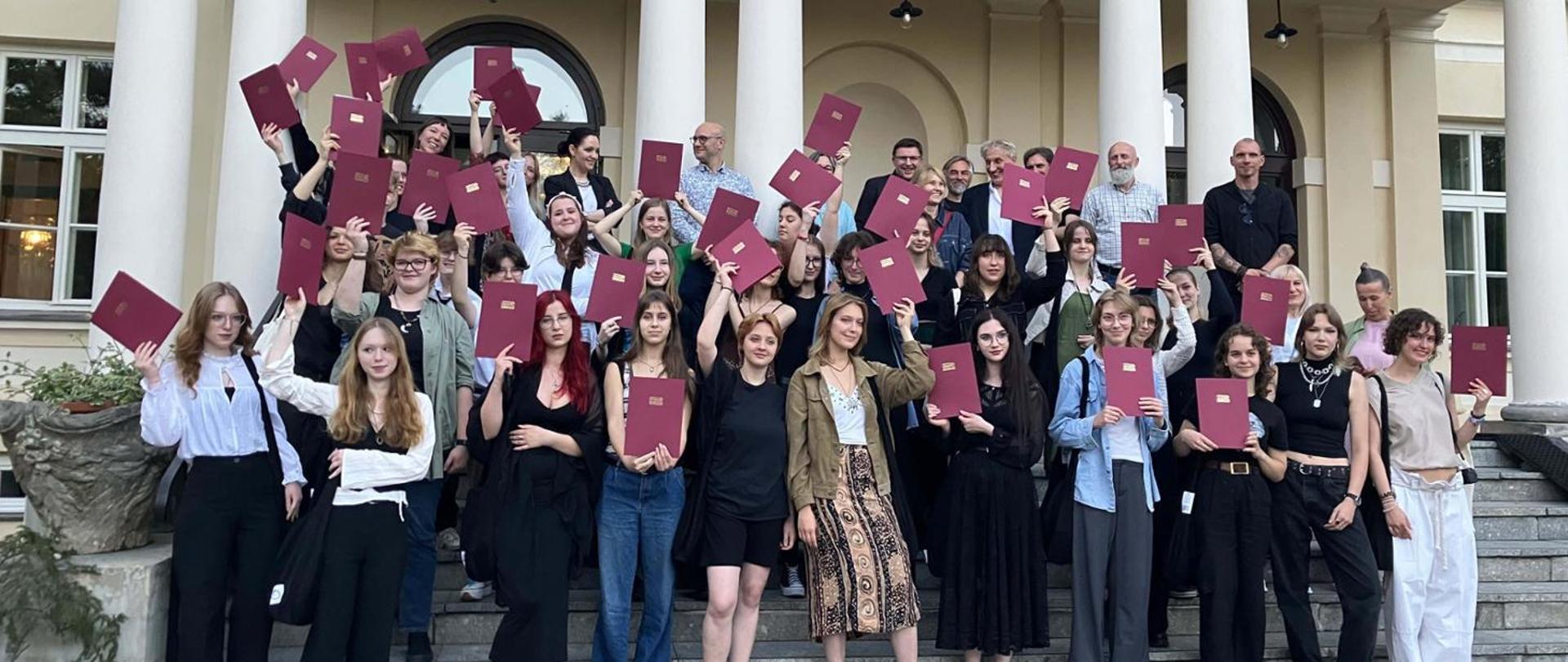 Uczestnicy konkursu, grupa młodzieży na schodach trzymająca w rękach uniesione teczki