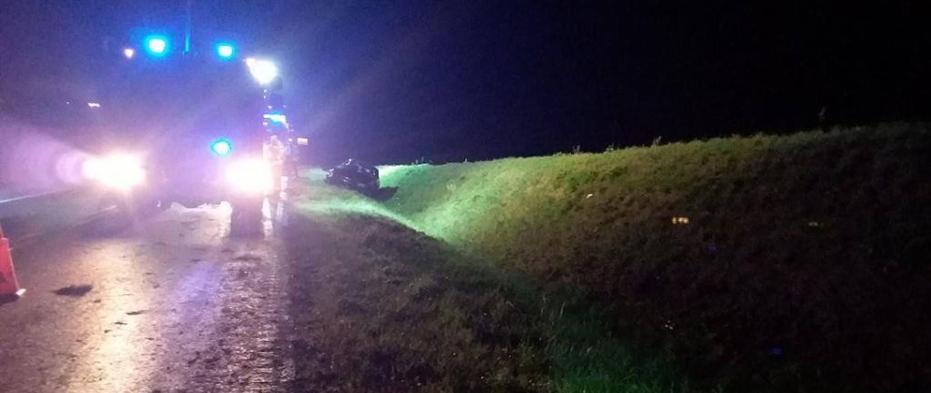 Pora nocna, oślepiające światła reflektorów samochodu strażackiego skierowane w obiektyw aparatu. Kontur samochodu strażackiego widoczny na skraju jezdni, w przydrożnym rowie kontur samochodu osobowego po wypadku. 