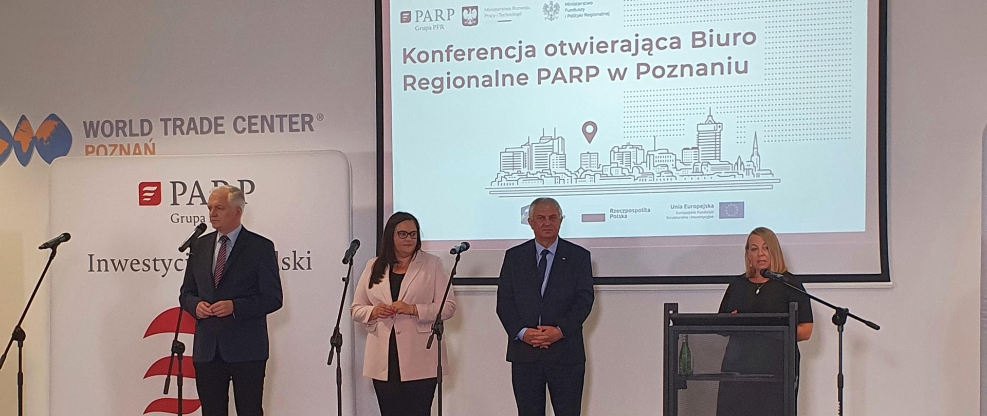 uczestnicy spotkania stoją na scenie, do mikrofonu mówi prezes PARP, za uczestnikami napis konferencja otwierająca Biuro Regionalne PARP w Poznaniu