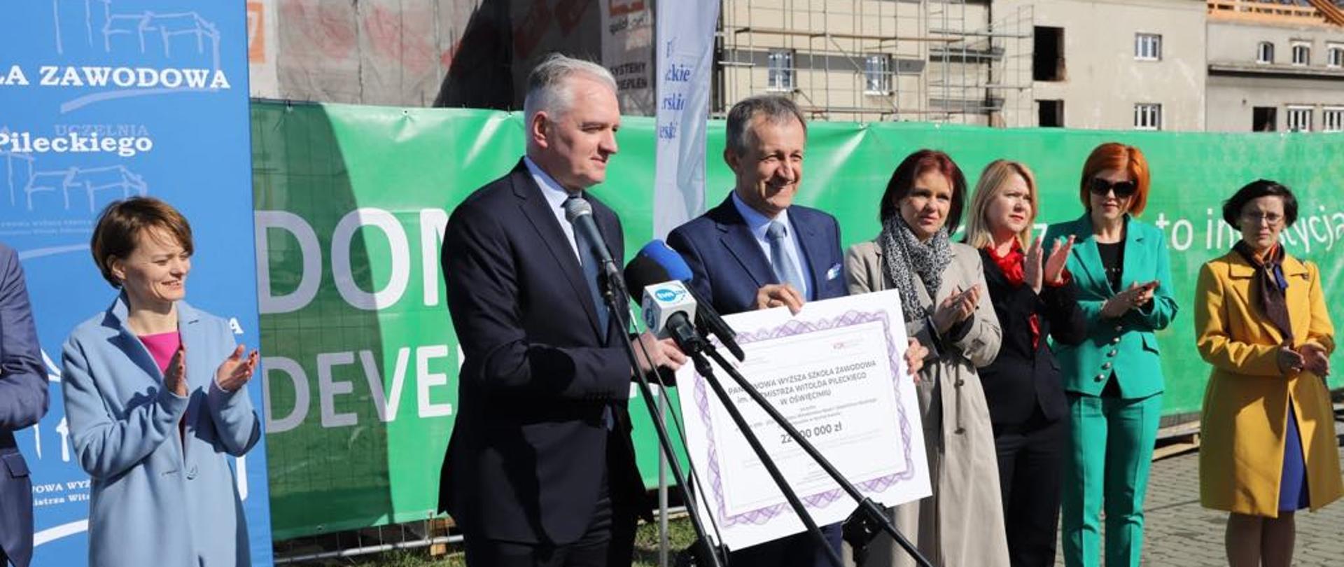 Minister Gowin przemawia do mikrofonu, w tle remontowane budynki