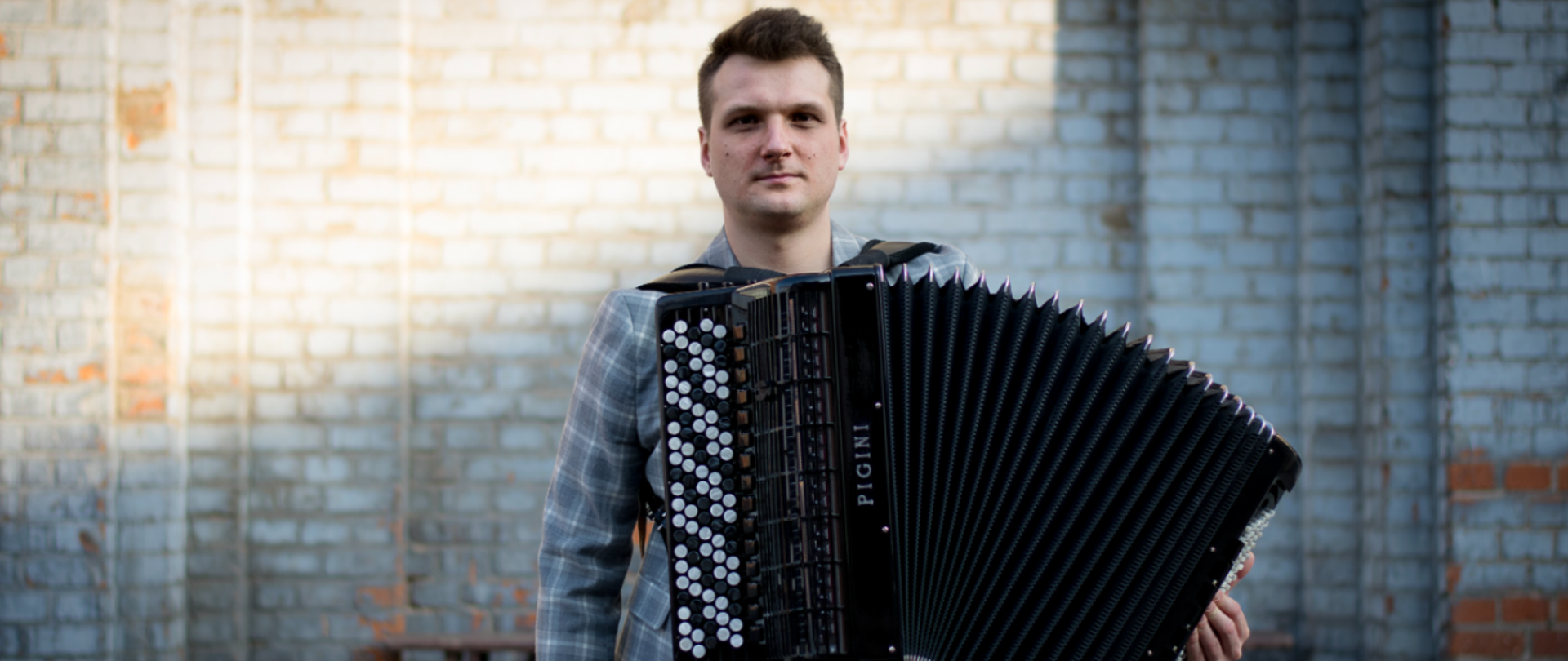 Mężczyzna ubrany w szarą kraciastą marynarkę, trzyma czarny akordeon guzikowy (miech instrumentu rozłożony w prawą stronę), stojąc na tle jasnej ceglanej ściany.