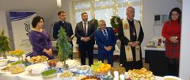 W centralnym miejscu zdjęcia znajduje się suto zastawiony stół na którym widać misy z kapustą z grochem, śledziami, sałatkami, patery z owocami i pasztecikami, sianko z opłatkiem oraz stroinki świąteczne. Za stołem, w centralnym miejscu stoi sześć osób. Od lewa: Zastępca Świętokrzyskiego Wojewódzkiego Inspektora Ochrony Roślin i Nasiennictwa- kobieta w fioletowej sukience, ksiądz w koloratce kapłańskiej, Wicewojewoda Świętokrzyski- mężczyzna w ciemnym garniturze oraz białej koszuli, Świętokrzyski Wojewódzki Inspektor Ochrony Roślin i Nasiennictwa w granatowym garniturze i tego samego koloru krawacie, Ksiądz Biskup oraz pracownik młoda kobieta ubrana na czerwono. Postaci stoją na tle okna oraz roll-upu z logo PIORiN. Wszyscy są zasłuchani w słowa przemawiającego biskupa.