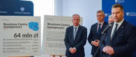 Minister Czarnek stoi i mówi do mikrofonu na stojaku, za nim stoi trzech mężczyzn w garniturach, obok nich na stojakach tablice z napisami Branżowe Centra Umiejętności i 64 mln zł, dalej biała ściana i zasłonięte pionowymi roletami okno.