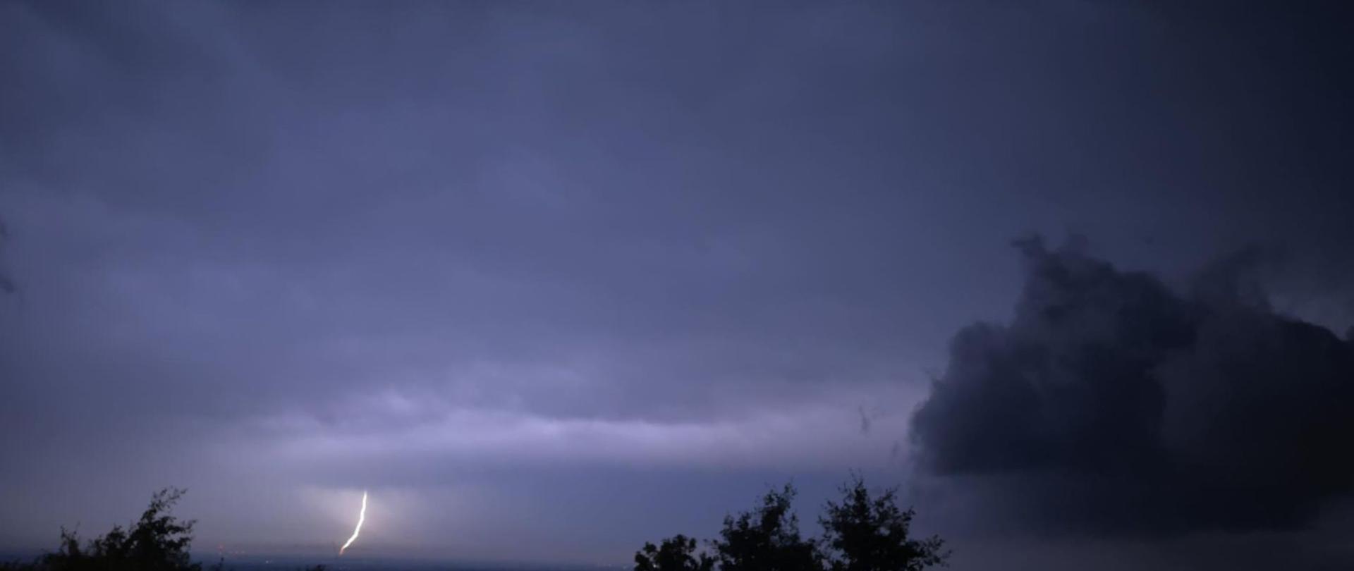 Zdjęcie przedstawia doziemienie piorunu na tle rozświetlonych chmur - zdjęcie wykonane w porze nocnej.