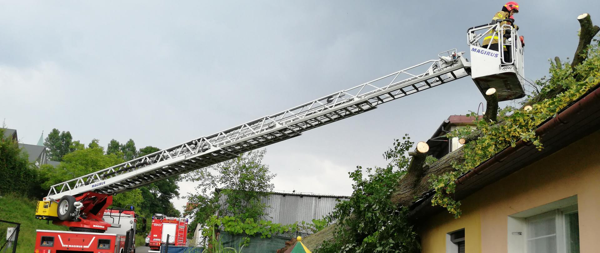 Na pierwszym planie po lewej stronie kadru stoi samochód z wysuniętą w prawą stronę kadru, drabiną mechaniczną. Na końcu drabiny w koszu ratowniczym, widać strażaka, przycinającego konary drzewa powalonego na budynek mieszkalny.