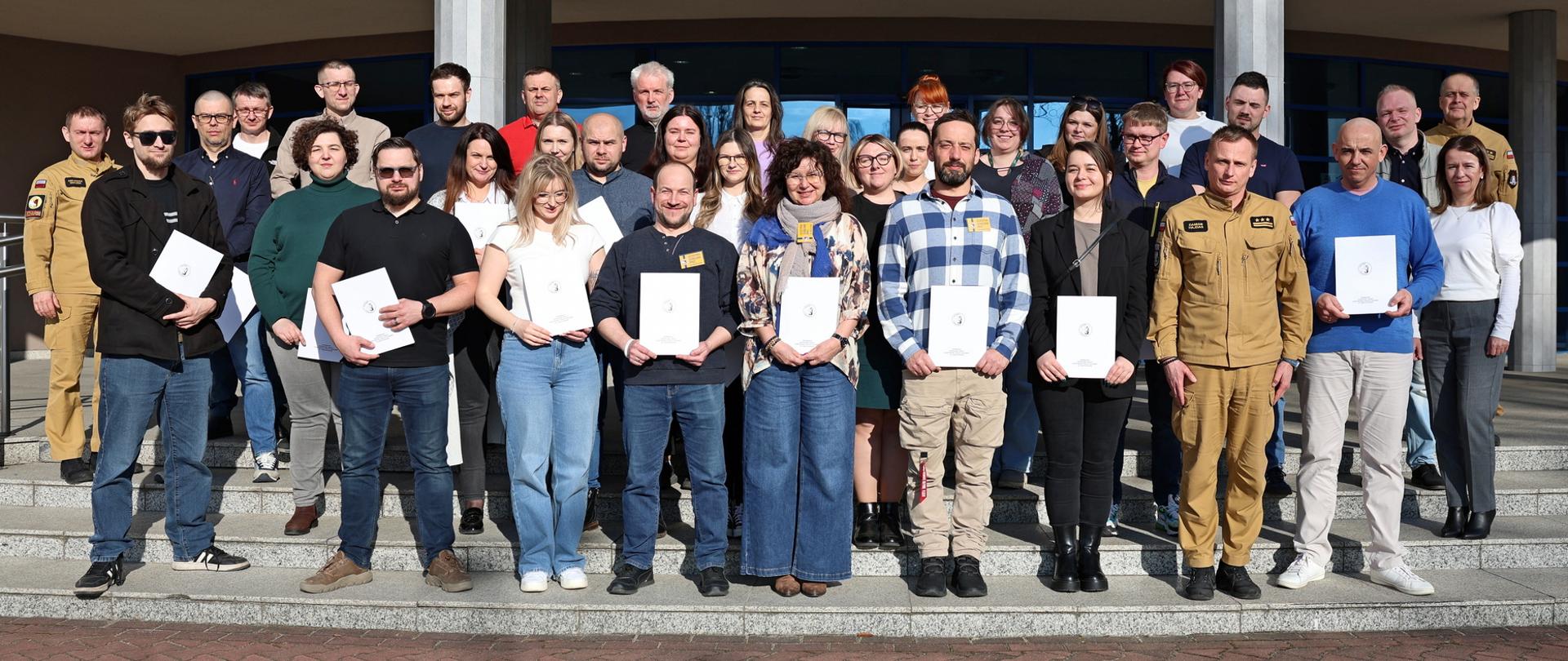 Pamiątkowe zdjęcie grupowe uczestników szkolenia inspektorów ochrony przeciwpożarowej (29 osób) w towarzystwie Zastępcy Komendanta Centralnej Szkoły PSP, kobiety oraz dwóch funkcjonariuszy CS PSP