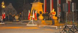 Narodowe Święto Niepodległości. Zdjęcie przedstawia pomnik Żołnierza Polskiego. Na cokole, na którym stoi pomnik, umieszczony jest orzeł. Za pomnikiem, na pionowych masztach zawieszone są 4 flagi biało-czerwone. Z kolei z prawej i lewej strony pomnika ustawione są dwa stojaki z flagami biało-czerwonymi. W tle budynki i drzewa. Na podwyższeniu przy mikrofonie, obok pomnika stoi strażak w umundurowaniu wyjściowym-zimowym. Obok pomnika stoją również uczniowie klasy mundurowej pełniący wartę honorową.