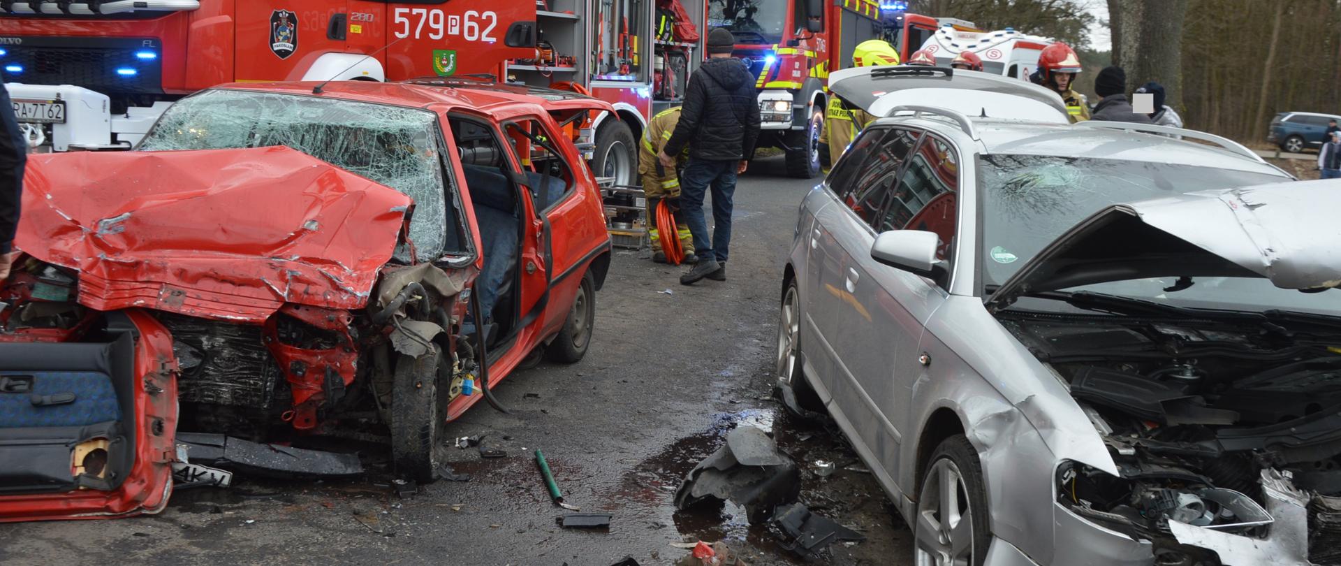 Czołowe zderzenie w Osieku Na jezdni i poboczu drogi stoją dwa rozbite pojazdy osobowe. Jeden jest srebrny, drugi - czrwony. Oba maja bardzo mocno uszkodzone przody. W tle wozy strażackie, karetka pogotowia i strażacy oraz las.