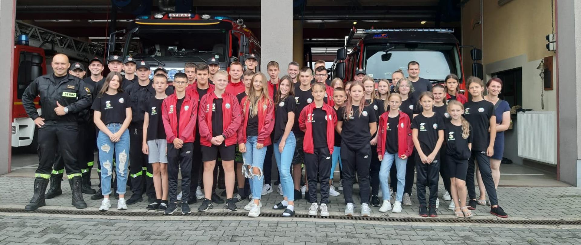 Grupowe zdjęcie uczestników obozu wraz z opiekunami oraz strażakami wykonane na tle samochodów pożarniczych stojących w garażu jednostki ratowniczo-gaśniczej CS PSP
