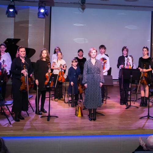 Zdjęcie przedstawia smyczkową orkiestrę kameralną szkoły pierwszego stopnia wraz z nauczycielami na scenie sali koncertowej przed wykonaniem utworu