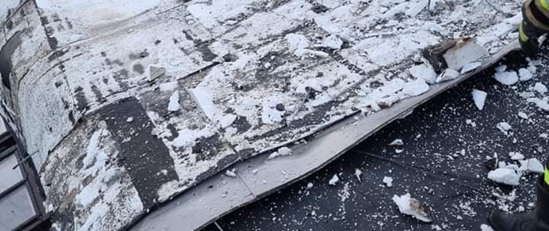 Na środku zdjęcia widać leżące na części dachu przekrycie stropodachu wykonane z papy i styropianu. Po prawej stronie zdjęcia widać dłoń i nogę strażaka , który usuwa uszkodzone elementy. W tle widoczna jest zielona ściana budynku , a po prawo od ściany widać gałęzie drzew.