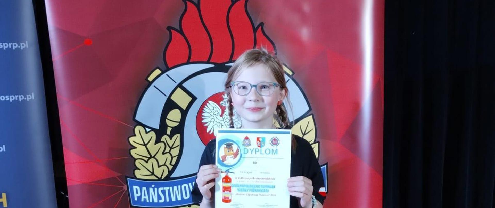 Zdjęcie przedstawia dziewczynek, która w ręku ma dyplom za zdobycie pierwszego miejsca OTWP.