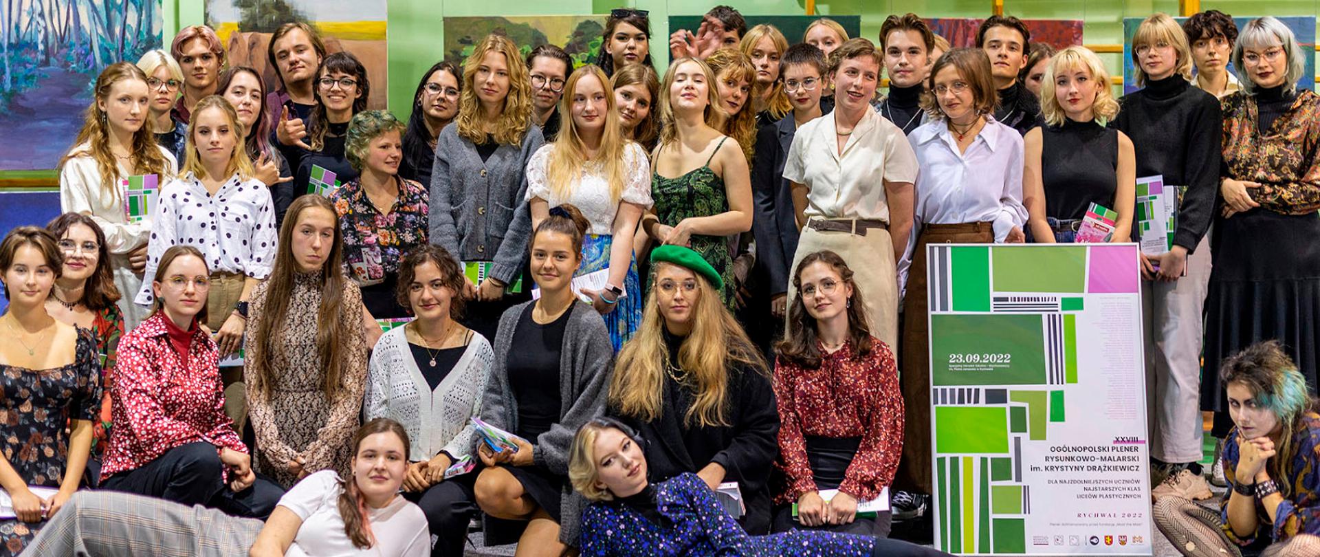 Zdjęcie zbiorowe 41 uczestników XXVIII Ogólnopolskiego Pleneru Rysunkowo-Malarskiego im. Krystyny Drążkiewicz dla najzdolniejszych uczniów najstarszych klas liceów plastycznych - Rychwał 2022 wykonane podczas wernisażu w Rychwale. W tle wiszące obrazy uczestników. W dolnej prawej części zdjęcia uczniowie trzymają plakat informacyjny o wernisażu.