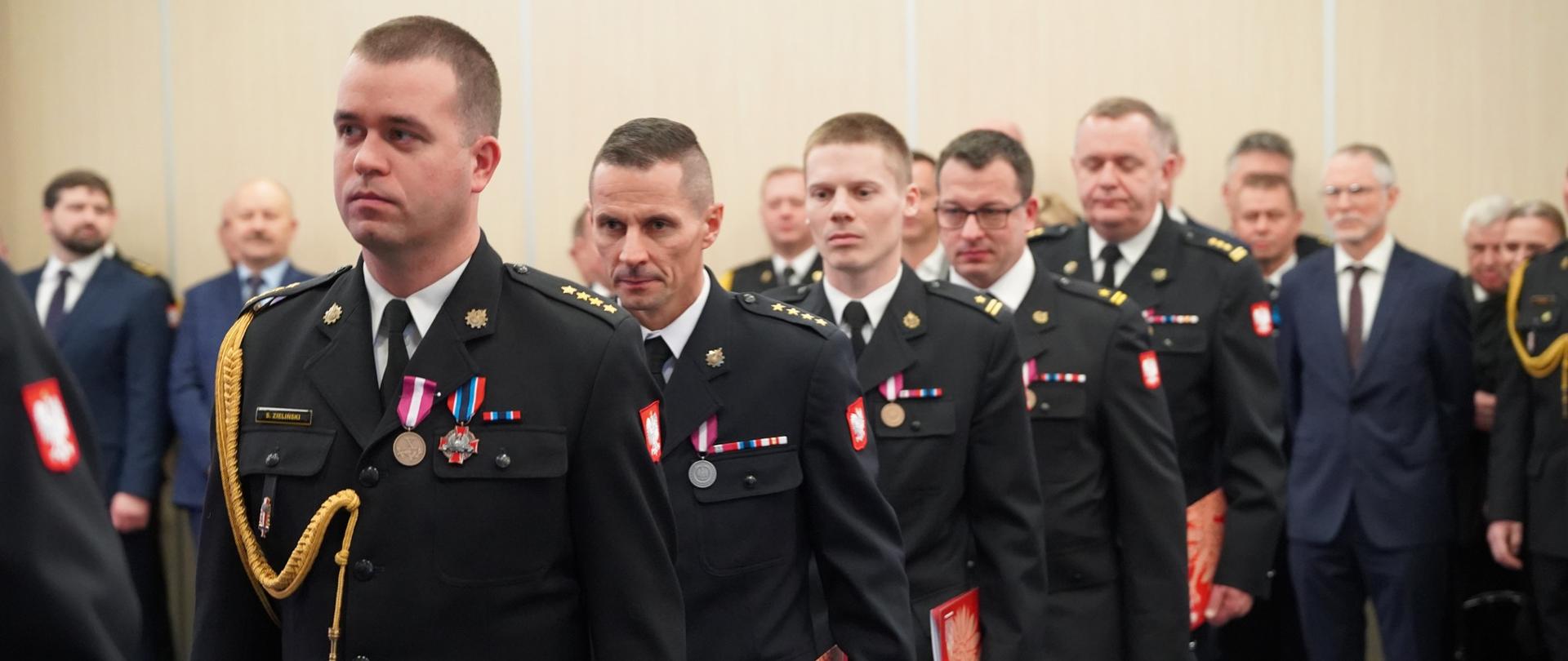 Strażacy odznaczeni medalami idą w szeregu, trzymając w rękach czerwone teczki z godłem
