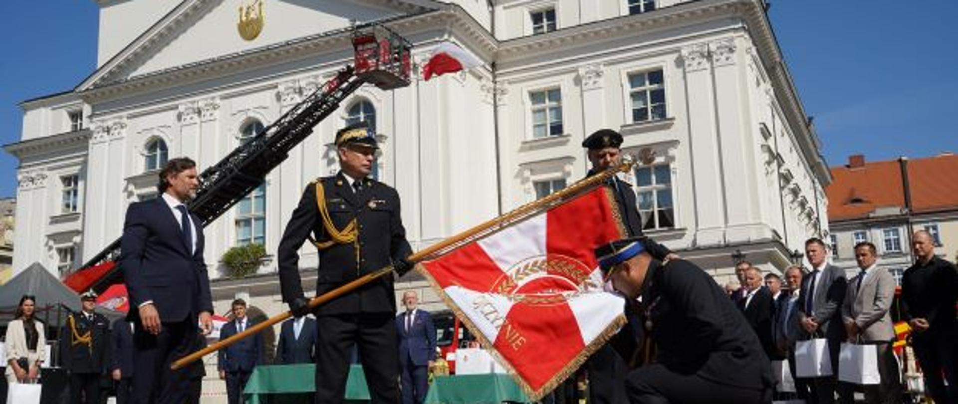 Na pierwszym planie widzimy dwóch mężczyzn strażaków ubranych w galowe ciemne mundury, czarne buty, czapki rogatywki oraz mężczyznę cywila ubranego w granatowy mundur. Jeden ze strażaków trzyma sztandar koloru biało-czerwonego i pochyla go w kierunku drugiego strażaka. Drugi strażak klęczy i całuje sztandar. Dalsza część planu to budynek ratusza okazały koloru białego. Zdarzenie obserwują zebrani goście również odświętnie ubrani w rękach trzymają torby z upominkami. 