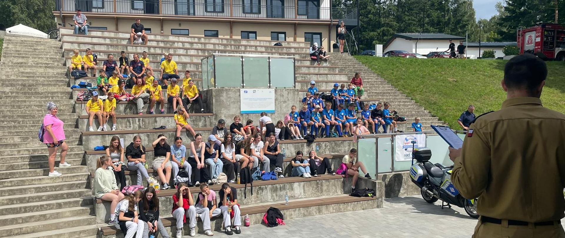 Zdjęcie przedstawia strażaka omawiającego wykonywane czynności podczas pokazu kwalifikowanej pierwszej pomocy. W tle widać dzieci i opiekunów.