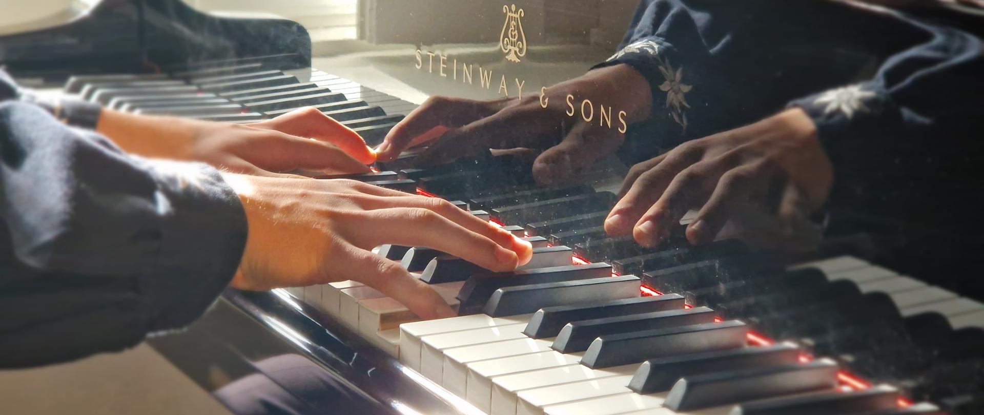Zdjęcie przedstawia zbliżenie na dłonie osoby grającej na czarnym, lakierowanym fortepianie marki Steinway & Sons. Instrument: Widoczna jest klawiatura oraz fragment obudowy fortepianu z wyraźnym, złotym logo producenta. Powierzchnia instrumentu jest tak czysta, że odbijają się w niej dłonie i rękawy grającego. Dłonie: Są ułożone na klawiszach w sposób sugerujący ruch i skupienie. Palce spoczywają zarówno na białych, jak i czarnych klawiszach. Oświetlenie: Zdjęcie charakteryzuje się silnym, ciepłym światłem (prawdopodobnie słonecznym), które wpada z boku, tworząc efektowne odblaski na krawędziach instrumentu i podkreślając fakturę skóry na dłoniach. Atmosfera: Całość ma elegancki, artystyczny i niemal intymny charakter, skupiając uwagę na kontakcie artysty z instrumentem.