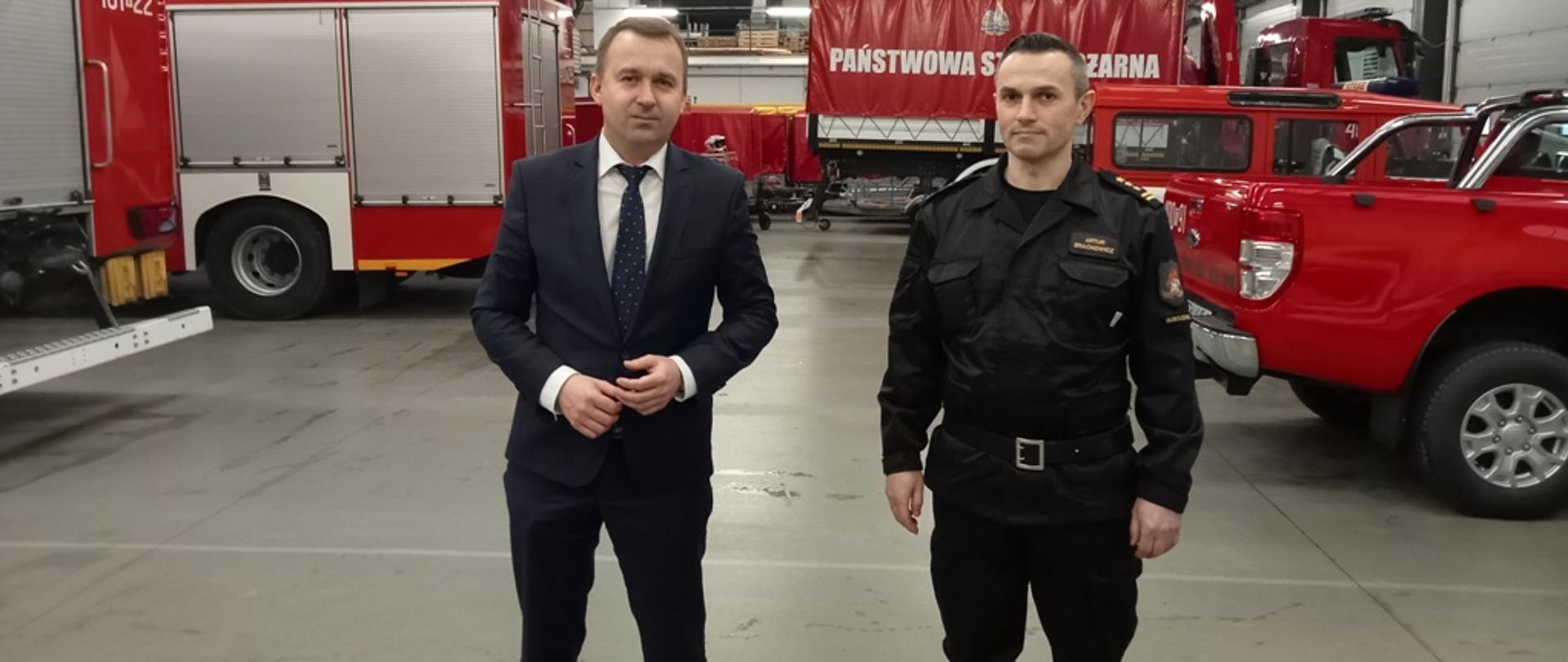 Zdjęcie przedstawia boksy garażowe Jednostki Ratowniczo-Gaśniczej w Busku-Zdroju.W centralnej części znajduje się Pan Minister Michał Cieślak wraz z komendantem powiatowym Państwowej Straży Pożarnej.W tle zdjęcia znajdują się pojazdy pożarnicze