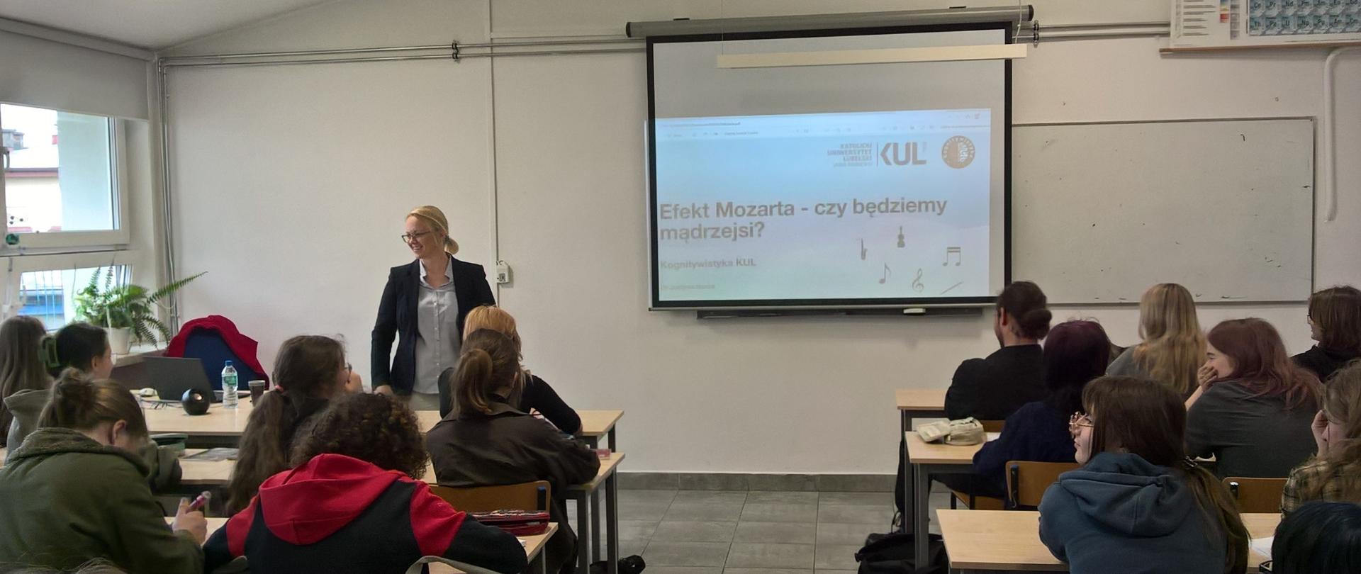 Zdjęcie przedstawia salę lekcyjną, w której odbywa się wykład lub prezentacja. Na pierwszym planie widzimy grupę uczniów lub studentów siedzących przy ławkach, zwróconych twarzą do prowadzącej. Prowadząca to kobieta w okularach, ubrana w jasną koszulę i ciemną marynarkę, stojąca obok ekranu projekcyjnego.
Na ekranie wyświetlony jest slajd z tytułem prezentacji: „Efekt Mozarta – czy będziemy mądrzejsi?”. W tle widać logotypy Katolickiego Uniwersytetu Lubelskiego Jana Pawła II (KUL) oraz napisy wskazujące na kierunek Kognitywistyka. Obok znajdują się ikony związane z muzyką, np. nuty.
Sala jest dobrze oświetlona, z dużym oknem po lewej stronie i rzutnikiem na suficie. Na ścianie po prawej stronie znajduje się tablica okresowa pierwiastków. Atmosfera na zdjęciu sugeruje skupienie i zainteresowanie uczestników.