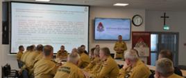 Strażacy województwa pomorskiego ubrani w mundury koloru piaskowego siedzą za stołami i patrzą na ekran multimedialny przed którym siedzi kadra kierownicza Komendy Wojewódzkiej Państwowej Straży Pożarnej w Gdańsku w liczbie czterech osób. 