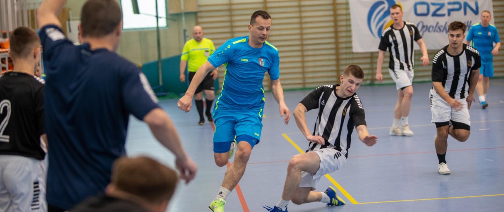 Zawodnik w czarno‑białym stroju wykonuje wślizg, próbując zatrzymać piłkę prowadzoną przez gracza w niebieskim stroju podczas turnieju eliminacyjnego PSP w futsalu w Kluczborku.