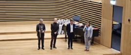 Trzech mężczyzn i kobieta stoją obok siebie na estradzie sali koncertowej. Jeden mężczyzna trzyma flet, kobieta przemawia do mikrofonu. Za nimi stoi fortepian i ułożone są nagrody w konkursie.