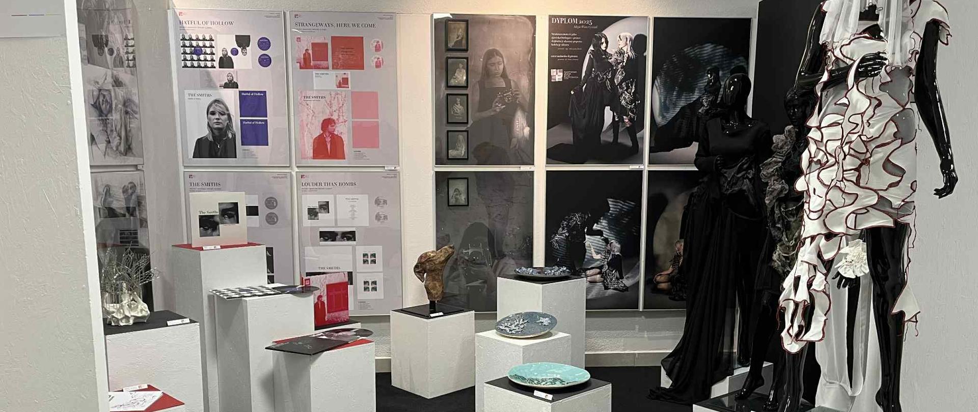 Na zdjęciu ekspozycja prac dyplomowych: ceramika - talerze - wyeksponowana na kubikach, grafika, prace z pracowni ubioru