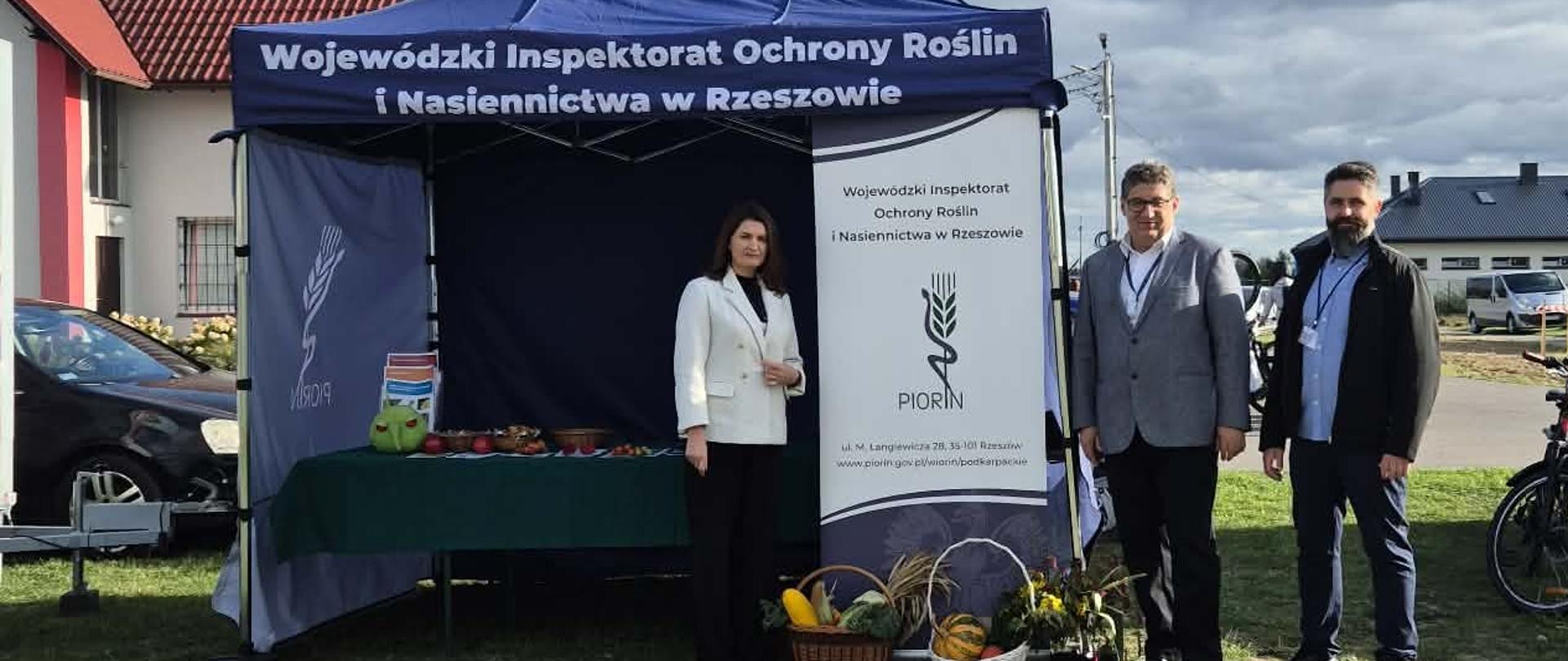 Stoisko informacyjne przy którym jest namiot z nazwą instytucji. Pod namiotem jest stolik na którym rozłożone są gadżety i ulotki, przy stoliku stoją osoby. 