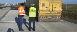 Umundurowany funkcjonariusz warmińsko-mazurskiej Inspekcji Transportu Drogowego stoi obok kierowcy z tyłu kontrolowanego zestawu ciężarowego.