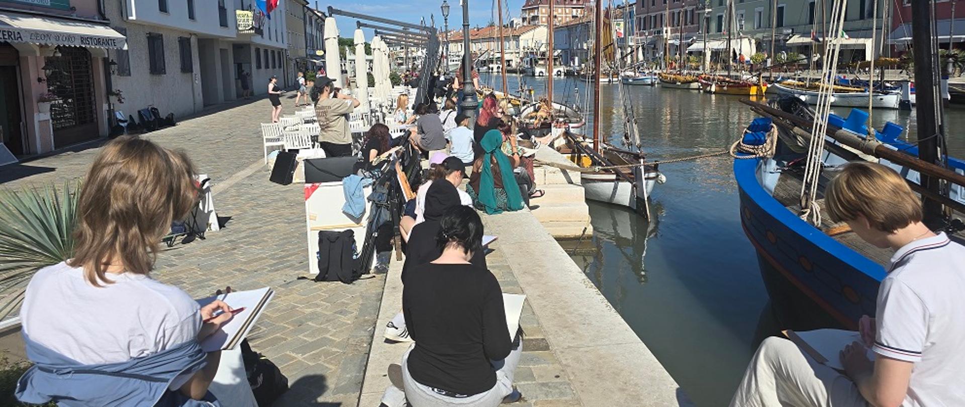 Na zdjęciu uczniowie podczas wykonywania prac malarskich. W tle kanał wypełniony wodą, żaglówki