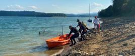 Przy brzegu jeziora pomarańczowa łódź, do której wsiadają płetwonurkowie. Dwóch z nich ma założone butle nurkowe. 