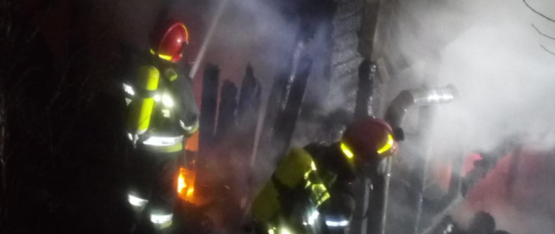 Zdjęcie przedstawia strażaków w ubraniach specjalnych, wykorzystujących sprzęt ochrony dróg oddechowych prowadzących działania gaśnicze. Podawany jest prąd gaśniczy wody. Widoczne są spalone elementy budynku.