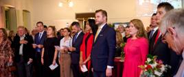 A foto mostra várias pessoas em traje de passeio completo, em pé, no interior de um salão histórico, com sembalntes sérias cantando o Hino da Polônia