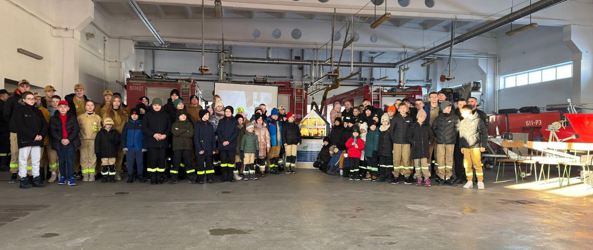 Pomieszczenie przypominające salę szkoleniową lub strażnicę, w której odbywa się prezentacja dotycząca bezpieczeństwa pożarowego i ekologii. Na pierwszym planie stoi mężczyzna w czarnym polarze z logo, prezentujący materiały edukacyjne.