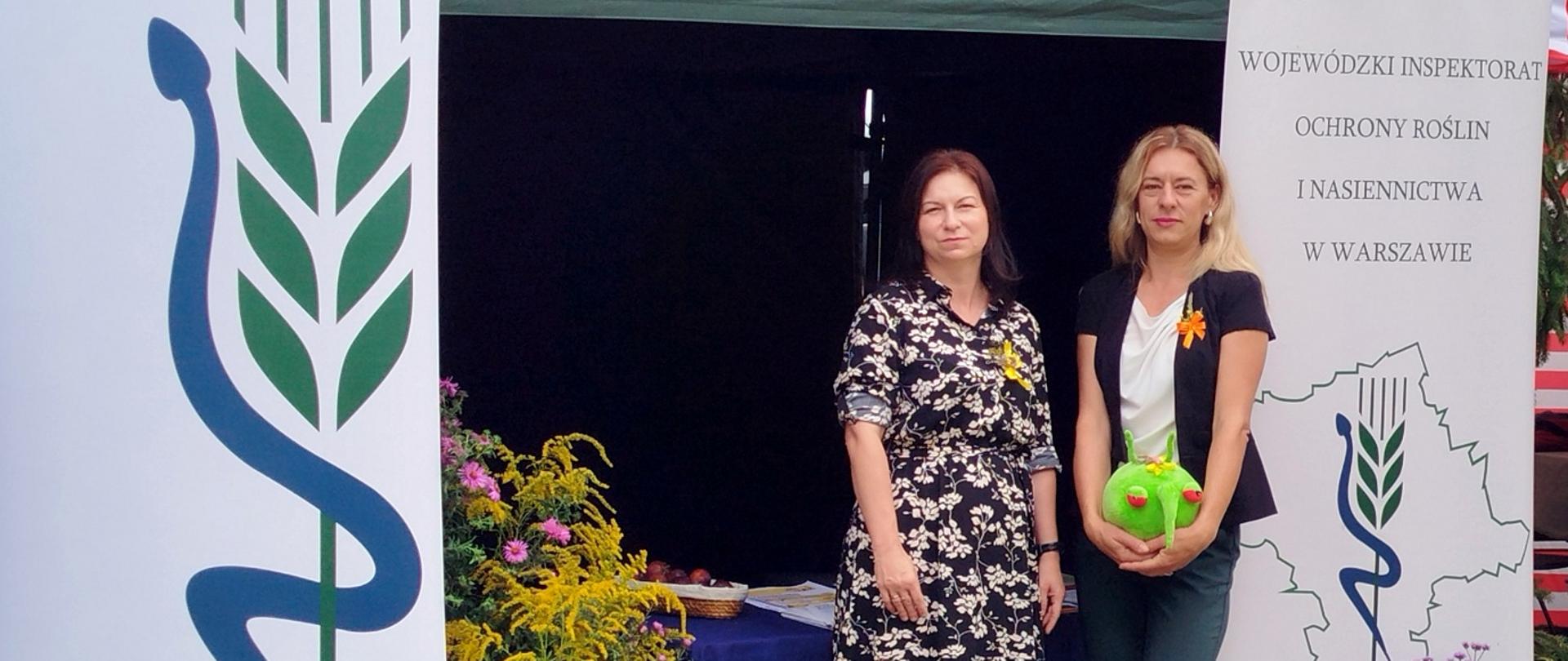 Stoisko plenerowe Wojewódzkiego Inspektoratu Ochrony Roślin i Nasiennictwa w Warszawie – pod zielonym namiotem stoją dwie kobiety, obok banery z logo PIORiN, a przed nimi kosze z kwiatami i warzywami.